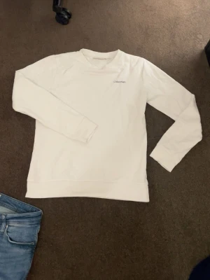 Vit sweatshirt från Calvin Klein - Snygg vit sweatshirt från Calvin Klein med diskret logga på bröstet. Tröjan har rund halsringning, långa ärmar och är gjord i mjukt material som känns skönt mot huden. Perfekt för en clean och stilren look.