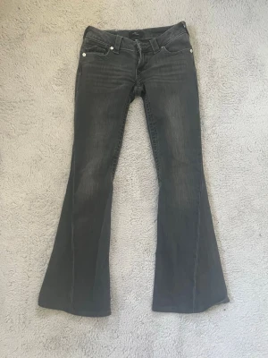 Grå bootcut jeans från True Religion - Säljer ett par grå bootcut jeans från True Religion med snygga detaljerade bakfickor och silverfärgade knappar. Jeansen har klassisk femficksdesign och är tillverkade i denim med lätt tvättad look. Perfekta för dig som gillar en avslappnad men trendig stil. W24 sydda så passar nån runt 160, skriv innan man köper 