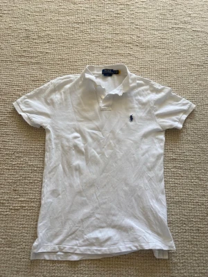 Vit piké från Polo Ralph Lauren - Klassisk vit pikétröja från Polo Ralph Lauren i slim fit-modell. Tröjan har korta ärmar, krage och knappar framtill samt den ikoniska broderade loggan på bröstet. Tillverkad i mjuk bomull som känns skön mot huden.