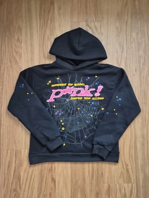 Svart sp5der hoodie med rosa tryck och stjärnor - Cool svart hoodie med stor huva och färgglatt tryck på ryggen. Trycket har texten 'Pink!' i rosa med vita prickar, gul text och ett stort spindelnätsmotiv. Dekorerad med små stjärnor i blått, gult och lila. Perfekt för dig som gillar unika och grafiska plagg.