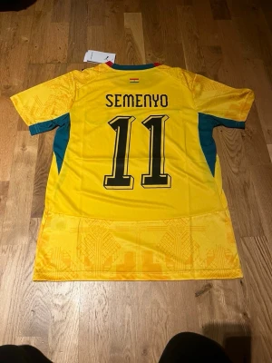 Ghana matchtröja Puma Semenyo #11 - Gul Ghana fotbollströja från Puma med Semenyo och nummer 11 på ryggen. Tröjan har svarta detaljer, landslagets emblem och stjärna på bröstet samt gröna inslag på sidorna. Tillverkad i lätt och ventilerande material, perfekt för match eller träning.