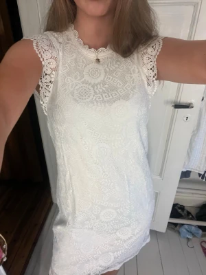 Vit spetsklänning  - Söt vit spetsklänning med vackra spetsdetaljer och blommigt mönster. Klänningen är ärmlös med dekorativa spetskanter vid axlarna och har en rak, kort passform. Perfekt för sommarens alla tillfällen och skolavslutning/student
