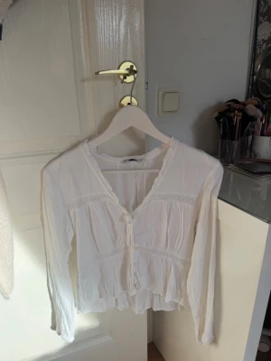 Vit romantisk blus med spetsdetaljer - Säljer en vit blus med v-ringning och dekorativa spetsdetaljer. Blusen har långa ärmar och knytning framtill, samt en lätt rynkad och flowy nederkant. Perfekt för en trendig och feminin look.