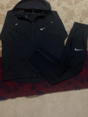 Svart Nike träningsset med huva - Säljer ett svart träningsset från Nike med jacka och matchande byxor. Jackan har huva och dragkedja, och både jacka och byxor har vita Nike-loggor. Perfekt för sport och träning, tillverkad i lätt och funktionellt material.