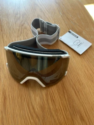 Salomon vita skidglasögon - Snygga vita skidglasögon från Salomon med bred, spegeltonad lins och justerbart elastiskt band. 