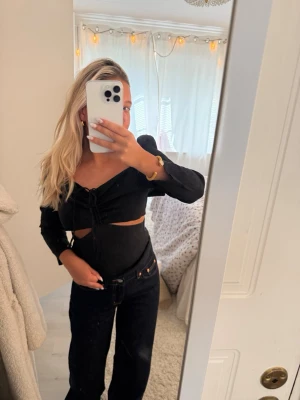 Svart croppad topp med snörning - Trendig svart croppad topp med långa ärmar och snygg snörning framtill. Toppen har cut-out detaljer vid midjan och en v-ringad urringning. Perfekt för dig som vill ha en edgy och stilren look.