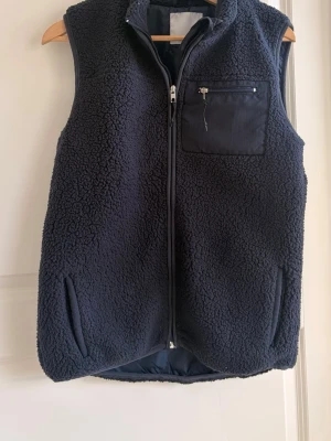 Mörkblå fleeceväst med dragkedja - Mysig mörkblå fleeceväst med hög krage och hel dragkedja framtill. Västen har en bröstficka med dragkedja och två sidofickor. Perfekt för lager-på-lager och chill stil. Den är i barnstorlek 160 men passar xs/s
