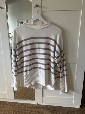 Vit och beige randig stickad tröja - Säljer en stickad tröja med breda beige och vita ränder. Tröjan har rund halsringning, långa ärmar och ribbstickade detaljer vid nederkant och ärmslut. Perfekt för att skapa en chill och trendig look. Köpt förra sommaren från MQ men märket är dobber. 