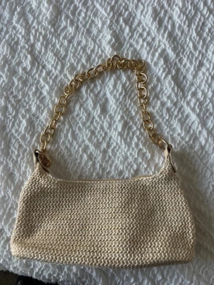 Beige stickad handväska med guldkedja - Snygg beige stickad handväska med en chunky guldfärgad kedja som axelband. Väskan har en avlång och mjuk form, perfekt för att bära det viktigaste. Materialet är stickat och ger en cool och trendig look.