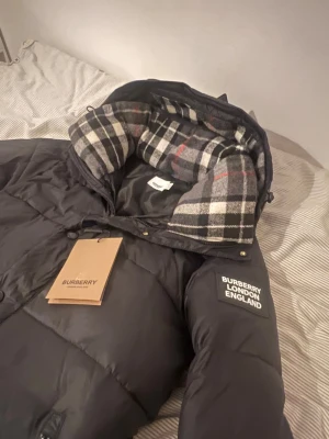 Svart dunjacka från Burberry - Snygg svart dunjacka från Burberry med klassiskt rutigt foder i huvan och tydlig Burberry London England-logga på ärmen. Jackan har dragkedja, knappar och två fickor framtill. Perfekt för kalla vinterdagar och riktigt stilren. Storlek M mem passar S. Går it göra t väst