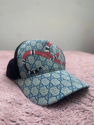 Zara keps med orm och GG-mönster - Snygg keps från Zara med klassiskt blått GG-monogram över hela framsidan och en röd orm som slingrar sig över kepsen. Baksidan är i svart mesh för extra ventilation. Perfekt för dig som vill sticka ut med en unik accessoar.