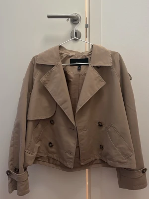 Kort beige trenchcoat från Vero Moda - Stilren kort trenchcoat i beige från Vero Moda. Jackan har dubbelknäppning, bred krage, axelklaffar och fickor fram. Tillverkad i ett mjukt bomullsliknande material och har justerbara ärmslut med knappar. Perfekt för en trendig och avslappnad look.