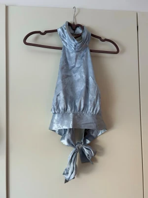 Ljusblå halterneck topp i satin - Snygg ljusblå halterneck topp i glansigt satinliknande material. Toppen har bred knytning i ryggen och hög krage som ger en cool och elegant look. Perfekt för dig som vill sticka ut med en unik topp.