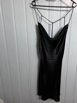 Svart satinklänning med tunna band - Säljer en svart satinklänning med tunna axelband och snygg draperad urringning. Klänningen har en slimmad passform och ett glansigt material som ger en lyxig känsla. Öppenrygg med dekorativa banddetaljer som gör den extra speciell.