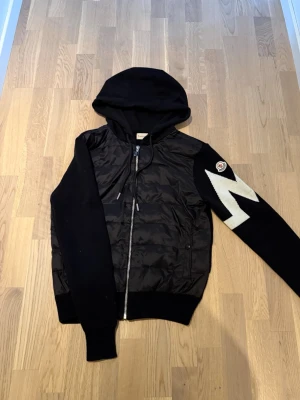 Svart Moncler hoodiejacka med mönster - Säljer en svart hoodiejacka från Moncler med dragkedja framtill och stickade ärmar. Ena ärmen har ett vitt zigzag-mönster och Moncler-logga. Jackan har huva och ribbade muddar. Perfekt för dig som vill ha en unik och snygg jacka.