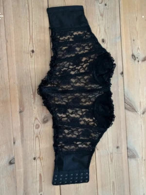 Svart shapewear med spetsdetaljer - Säljer en svart shapewear med hög midja och snygga spetsdetaljer. Modellen har hakknäppning nertill och är tillverkad i elastiskt material för en smickrande passform. Perfekt för att forma midjan och ge en snygg siluett.