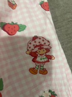 Rosa pyjamasbyxor med strawberry shortcake tryck - Säljer ett par rosa pyjamasbyxor med rutigt mönster och gulligt jordgubbstryck samt figurer från Strawberry Shortcake. Byxorna har resår och dragsko i midjan för skön passform. Perfekta för mysiga hemmakvällar!