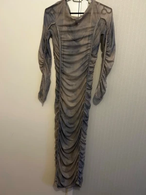 Grå långklänning med rynkade detaljer - Säljer en grå långklänning med rynkade detaljer längs hela framsidan och ärmarna. Klänningen har lång ärm och en kroppsnära passform som framhäver siluetten. Materialet är tunt och stretchigt, perfekt för en trendig look.