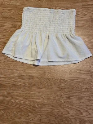 Vit axelbandslös bandeau topp - Säljer en söt vit bandeau topp med smockad överdel och volang nedtill. Toppen är axelbandslös och har en luftig, flowy passform som passar perfekt till sommaren. Perfekt att matcha med jeans eller kjol för en fräsch look.