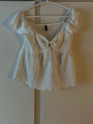 Vit blus med knyt och volang - Söt vit blus från H&M med korta volangärmar och knyt framtill. Toppen har en lätt rynkad struktur och v-ringad hals. Perfekt för en somrig look och passar till både jeans och kjol.