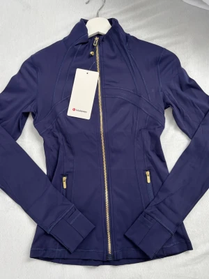 Marinblå träningsjacka från Lululemon - Snygg marinblå träningsjacka från Lululemon med guldfärgade dragkedjor och figurnära passform. Jackan har långa ärmar och två fickor med dragkedja. Perfekt för gymmet eller löprundan. Helt oanvänd med prislapp pågrudn av att jag fick den i present och den var för liten. STORLEK 4 påse tillkommer också