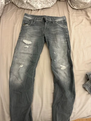 Grå slitna jeans från G-Star - Säljer ett par grå jeans från G-Star med slitna detaljer och distressed look. Jeansen har raka ben, klassisk femficksmodell och tydliga slitningar på låren och knäna. Materialet är denim i bomull och de har en cool, avslappnad vibe.