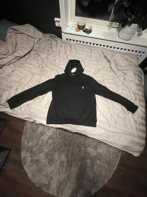 Svart hoodie från Polo Ralph Lauren - Snygg svart hoodie från Polo Ralph Lauren med klassisk huva och känguruficka framtill. Ikoniska vita Polo-loggan broderad på bröstet. Tillverkad i mjukt material som känns skönt mot huden. Perfekt för en avslappnad och stilren look. Passar om man är runt 170 cm lång, använd någon enstaka gång, inga fläckar eller skador.