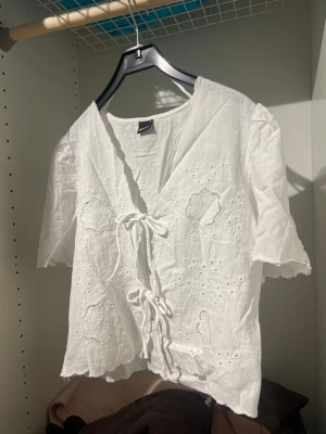 Vit broderad blus med knytning - Söt vit blus från Monki med broderade blommor och två knytband framtill. Blusen har korta puffärmar och vågig kant nertill. Tillverkad i ett lätt och luftigt tyg, perfekt för varma dagar. Romantisk och trendig look med fina detaljer.