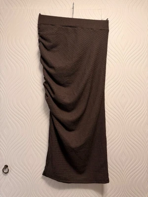 Brun långkjol med drapering - Säljer en brun långkjol med snygg drapering på sidan. Kjolen har en rak passform och är tillverkad i ett mjukt, strukturerat tyg som ger en cool look. Perfekt att styla med en enkel topp eller stickad tröja.