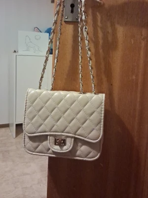 Beige quiltad axelväska med kedja - Snygg beige axelväska med quiltat mönster och kedjerem i metall och konstläder. Väskan har ett vridlås i silverfärg och en klassisk, boxig form. Perfekt för dig som gillar stilrena accessoarer med en trendig touch.