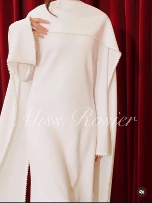 Vit långklänning från Miss Rosier - Elegant vit långklänning från Miss Rosier med slits och dekorativa pärldetaljer längs kanterna. Klänningen har lång ärm och en svepande, cape-liknande design som ger ett exklusivt intryck. Perfekt för dig som vill sticka ut med en unik och sofistikerad look.