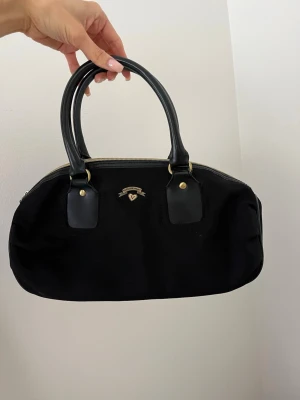Victorias secret bag med gulddetaljer - Stilren svart handväska i tyg med dubbla handtag i konstläder och guldfärgade metalldetaljer. Väskan har en oval form och dragkedja upptill. 