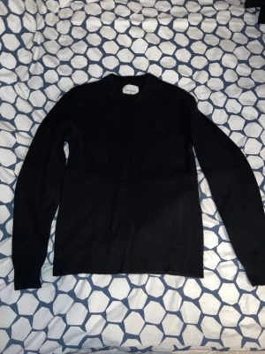 Svart stickad tröja ARKET - En stilren svart stickad tröja från ARKET med rund halsringning och långa ärmar. Tröjan har ribbade muddar vid ärmslut och nederkant. Perfekt basplagg i garderoben och enkel att matcha med olika outfits. 