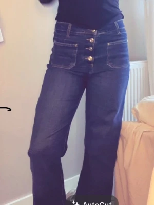 Mörkblå high waist jeans med knappar - Säljer ett par mörkblå high waist jeans med raka ben och fyra guldknappar framtill. Jeansen har stora fickor fram och bak, samt kontrastsömmar. Perfekta för dig som gillar en retro och trendig look. Klippta ner till