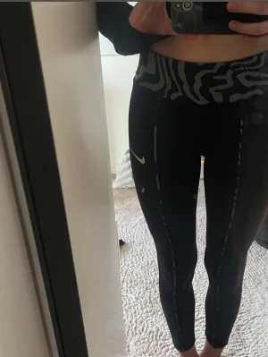 Nike svarta leggings med zebramönster - Säljer ett par svarta leggings från Nike med högt zebramönstrat midjeband och gul logga på benet. Stretchigt material som sitter tight och har dekorativa sömmar längs benen. Perfekta för träning eller chill dagar. En rosa fläck p knät syns inte men det är en rosa fläck men det är färg så går inte ta bort men man ser inte den så bra ifall man kolla långt ifrån