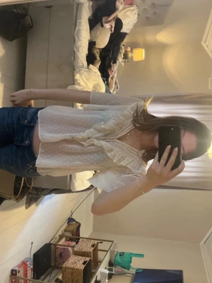 Vit  blus med volang - Söt vit blus med små prickar och volangdetaljer längs V-ringningen. Blusen har korta ärmar och ett lätt, luftigt tyg som ger en romantisk känsla. Perfekt att styla med jeans eller kjol för en trendig look.