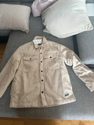 Beige overshirt i manchester från Zara - Säljer en beige overshirt i manchestertyg från Zara. Skjortjackan har två stora bröstfickor med lock och svarta knappar framtill. Perfekt för lager på lager och ger en chill vibe till din outfit.