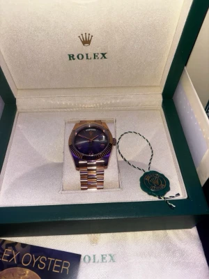  Oyster klocka i guld - Exklusiv  Oyster klocka med guldfärgat armband och mörkblå urtavla. Klockan har klassisk rund form och levereras i originalbox med certifikat och tillbehör. Perfekt för dig som vill ha en lyxig accessoar med ikonisk design.