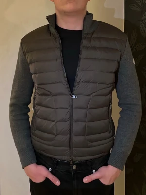 Mörkgrå dunväst från Moncler - Snygg mörkgrå dunväst från Moncler med quiltad front och två dragkedjeförsedda fickor. Ärmar och rygg i mjukt, stretchigt tyg för extra komfort. Klassisk Moncler-logga på vänster ärm och hög krage för en clean look.