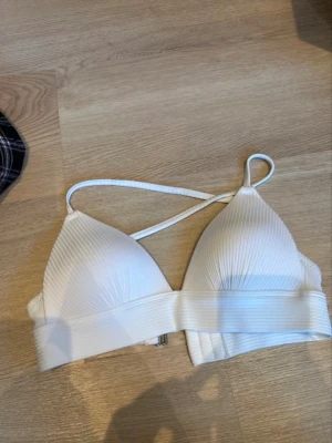 Vit ribbad bikiniöverdel från H&M - Säljer en vit bikiniöverdel från H&M med ribbad struktur och triangelformade kupor. Den har smala axelband som korsas i ryggen och bred resår under bysten för extra stöd. Metallspänne bak för enkel stängning. Perfekt för strand och pool.