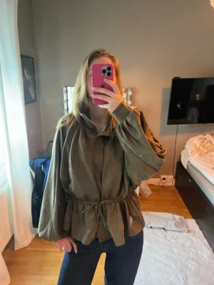 Khaki färgad vårjacka med snörning i midjan - Säljer en khaki färgad jacka med snörning i midjan och vida ärmar. Jackan har en avslappnad passform och är tillverkad i ett mjukt material som påminner om mocka. Den är ALDRIG använd och helt ny! 