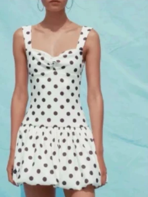 Vit prickig klänning - Söt vit kortklänning med svarta prickar och breda axelband. Klänningen har en figurnära överdel och en voluminös kjol med volang. Perfekt för sommarens alla tillfällen.