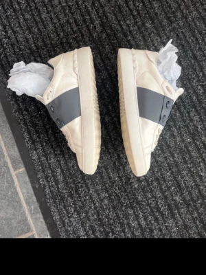 Vita Valentino sneakers med grå detalj - Snygga vita sneakers från Valentino med bred grå rem över snörningen och diskret logga på plösen. Skorna har lågt skaft och klassisk siluett. Tillverkade i läder med gummisula och coola nitar på hälen för extra edge.