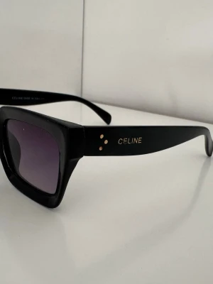 Svarta solglasögon Celine - Snygga svarta solglasögon med rektangulära bågar och mörka tonade glas. Klassisk och stilren design med Celine-logga i guld på sidan. Bågarna är i blankt syntetmaterial och ger en cool vibe till din look.