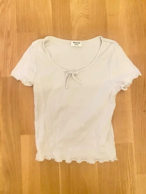 Vit ribbad t-shirt med rosett Kappahl - Söt vit ribbad t-shirt från Kappahl med korta ärmar och liten rosett framtill. T-shirten har vågiga kanter vid ärmslut och nederkant samt en mjuk, stretchig känsla. Perfekt för en enkel och stilren look.