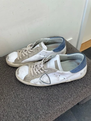 Philippe model skor storlek 46 - Snygga vita sneakers från Philippe Model med beige mockadetaljer och blå häl. Skorna har klassisk låg profil, vita snören och en ikonisk sköldlogga på sidan. Ovandelen är i läder och mocka, vilket ger en schysst mix av material och stil.