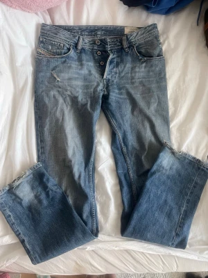 Blå bootcut jeans från Diesel - Säljer ett par blå bootcut jeans från Diesel med slitningar och knappar i gylfen. Jeansen har klassisk femficksmodell och coola detaljer som ger en avslappnad vibe. Perfekta för dig som gillar en lite mer relaxed och vintage look.
