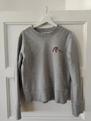 Grå sweatshirt från Replay - Klassisk grå sweatshirt från Replay med rund halsringning och broderad logga i rött och svart på bröstet. Tröjan har långa ärmar och ribbade muddar vid ärmslut och nederkant. Perfekt för en avslappnad och stilren look. Passar som M