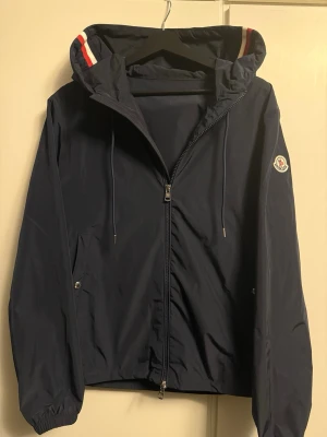 Blå vindjacka från moncler (med kvitto) - Säljer en mörkblå vindjacka frän Moncler med huva och dragkedja. Jackan har Moncler-logga pa ärmen och snygga rödvita detaljer på huvan. Pertekt för blåsiga dagar och enkel att matcha med det mesta. Den är i nytt skick, använd ett fåtal gånger. Säljs då den inte kommer till användning. Storlek 2, passar S