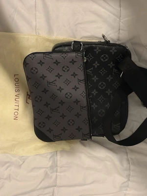 axelväska i svart/grå - Snygg axelväska i svart och grått med klassiskt monogrammönster. Väskan har justerbar axelrem, två fack med dragkedja och detaljer i silverfärg. Tillverkad i slitstark canvas med exklusiv känsla. Perfekt för dig som vill ha en trendig och praktisk väska.